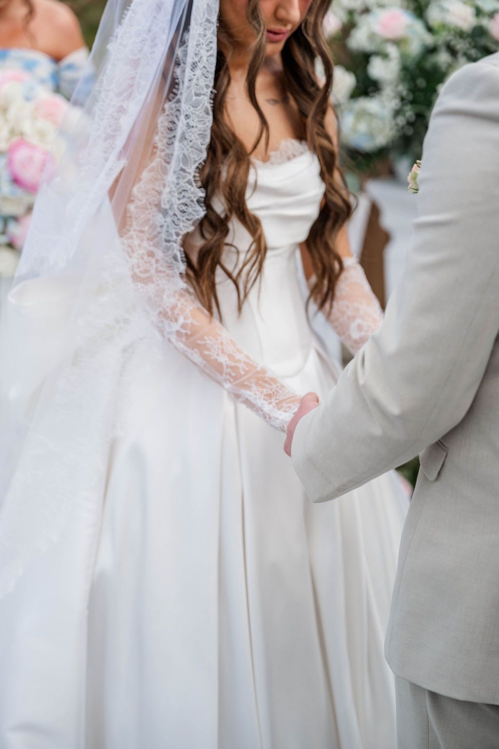 Bride in white lace gown holding hands with groom in light gray suit at a wedding ceremony