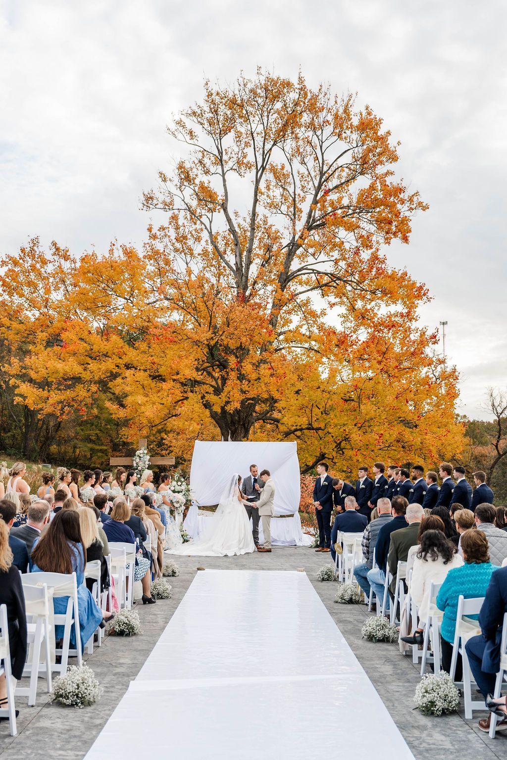 Outdoor wedding ceremony with guests seated on a white aisle under orange autumn trees