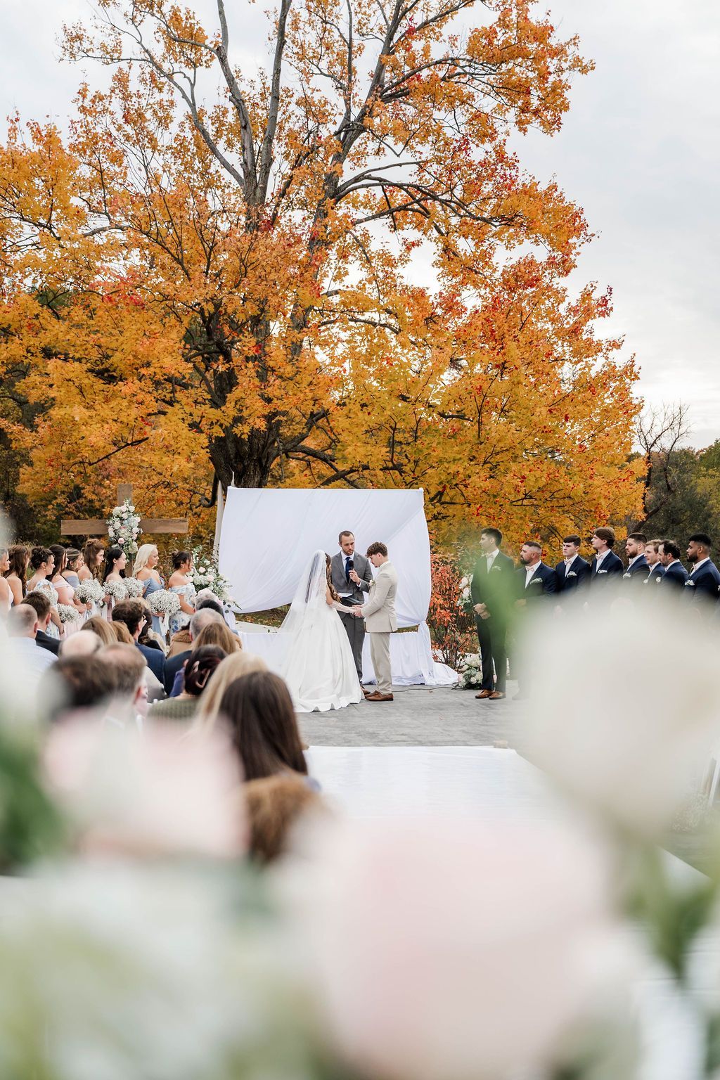 Outdoor wedding ceremony under orange autumn trees with bride, groom, and officiant at the altar