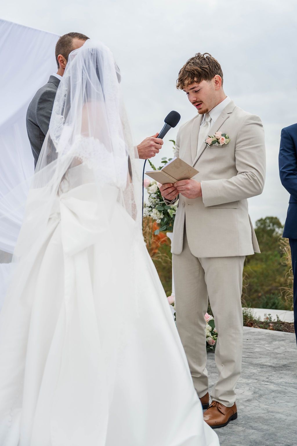 Bride in white veil and groom in beige suit exchange vows outdoors at a wedding ceremony.