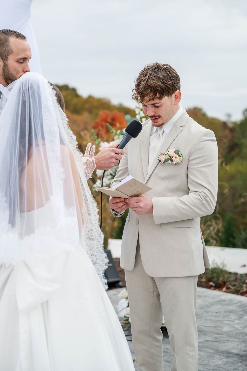 Bride and groom exchanging vows outdoors, with the groom reading from a paper as the bride holds a microphone.