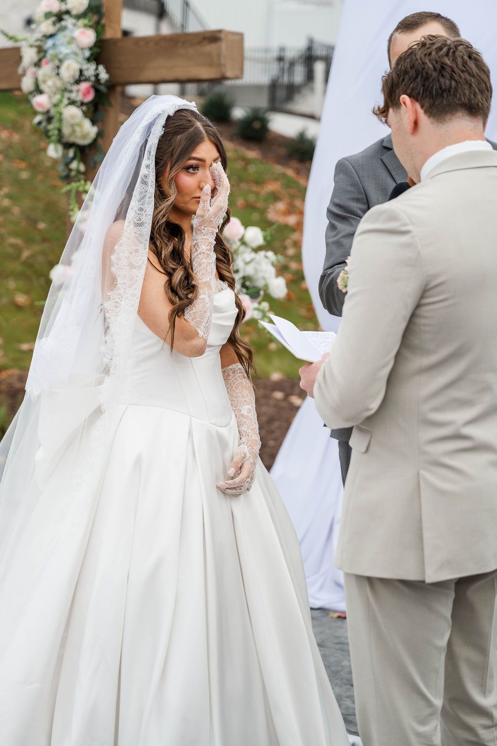 Bride in white dress and veil during outdoor wedding ceremony, facing the groom and officiant
