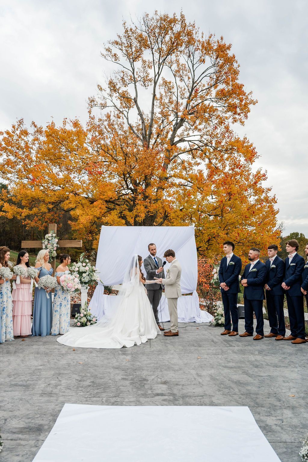Wedding ceremony outdoors beneath autumn trees, with bride and groom standing at the altar surrounded by attendants.
