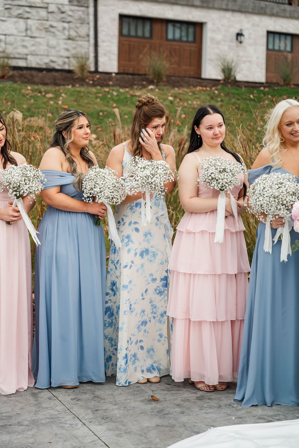 Bridesmaids in pastel dresses holding white bouquets stand outdoors in front of a stone building.