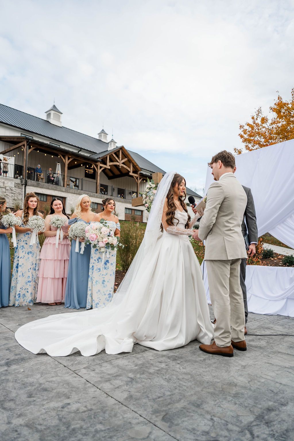 Bride and groom face each other at an outdoor wedding ceremony with bridesmaids in pastel dresses nearby.