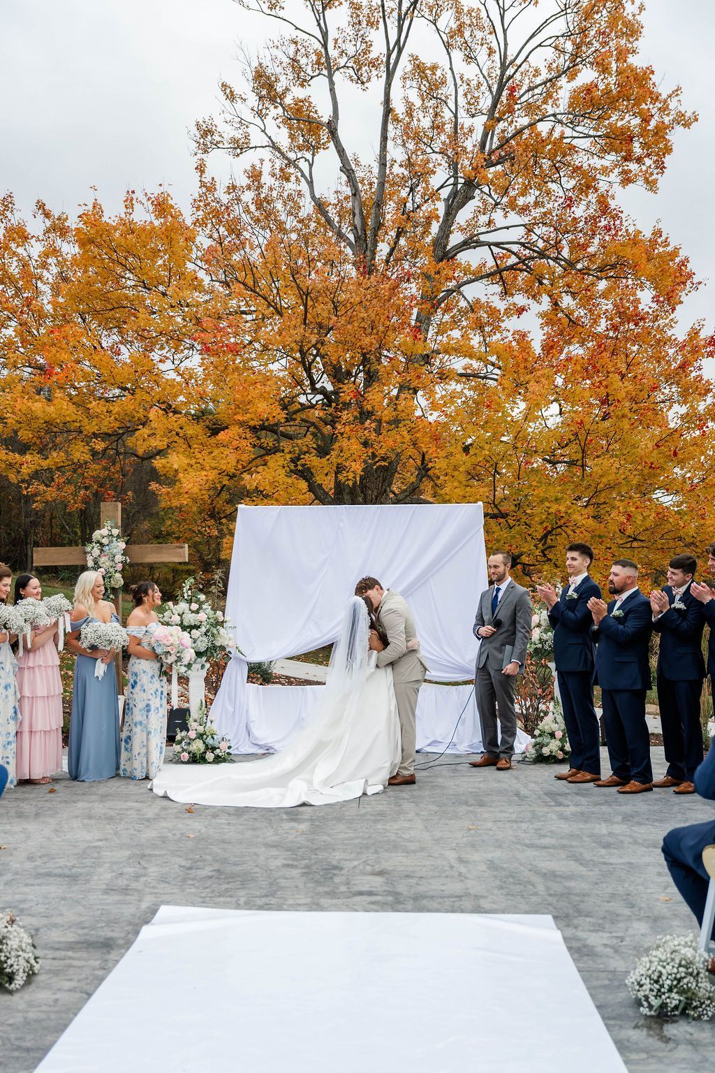 Outdoor wedding ceremony with bride and groom under autumn trees, guests standing on both sides