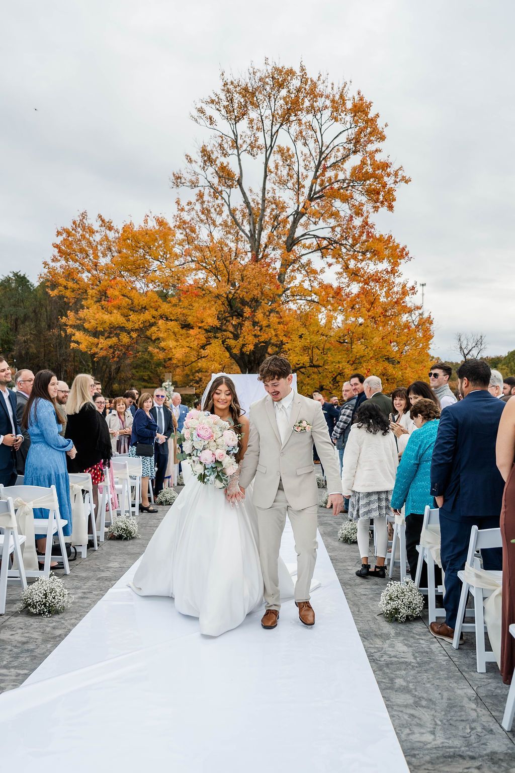 Newlyweds walking down an outdoor aisle lined with guests under autumn trees
