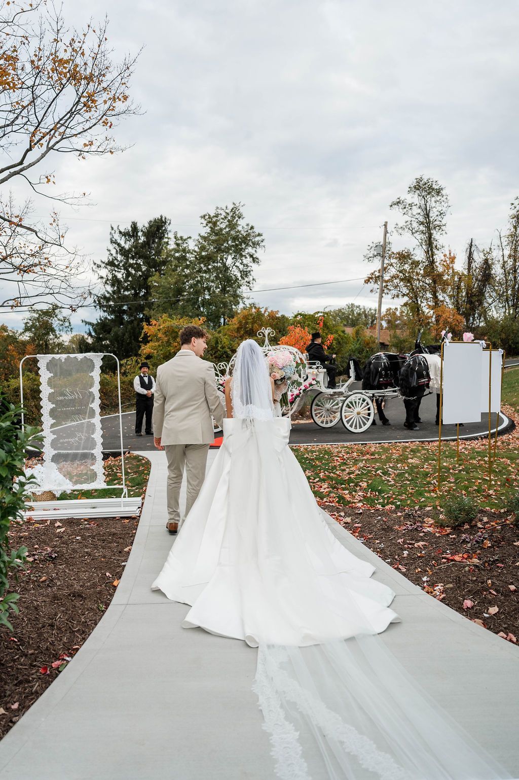 Bride and groom walking down an outdoor path at a wedding ceremony with autumn foliage and guests ahead