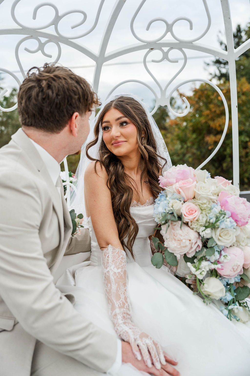 Couple seated in a white gazebo, bride holding a pastel bouquet and smiling at the groom.