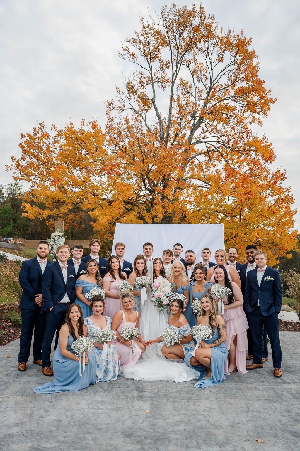 Large wedding party posing outdoors in formal attire before a white backdrop and autumn trees