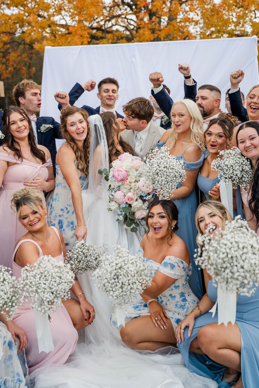 Wedding party posing outdoors with bouquets, bridesmaids in pastel dresses and groomsmen behind a white backdrop