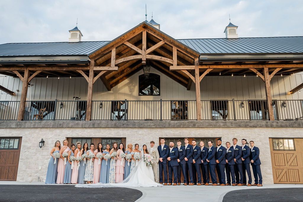 Large wedding party posing in front of a rustic barn-style venue with a covered porch and stone facade
