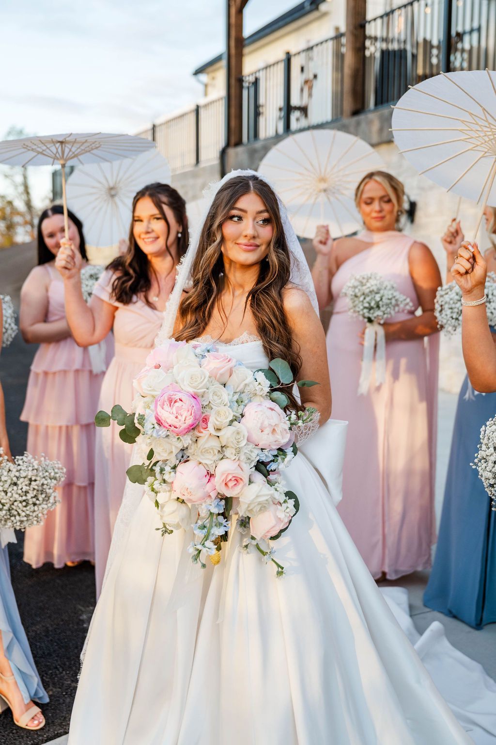 Bride holding pastel bouquet with bridesmaids behind her holding parasols outdoors