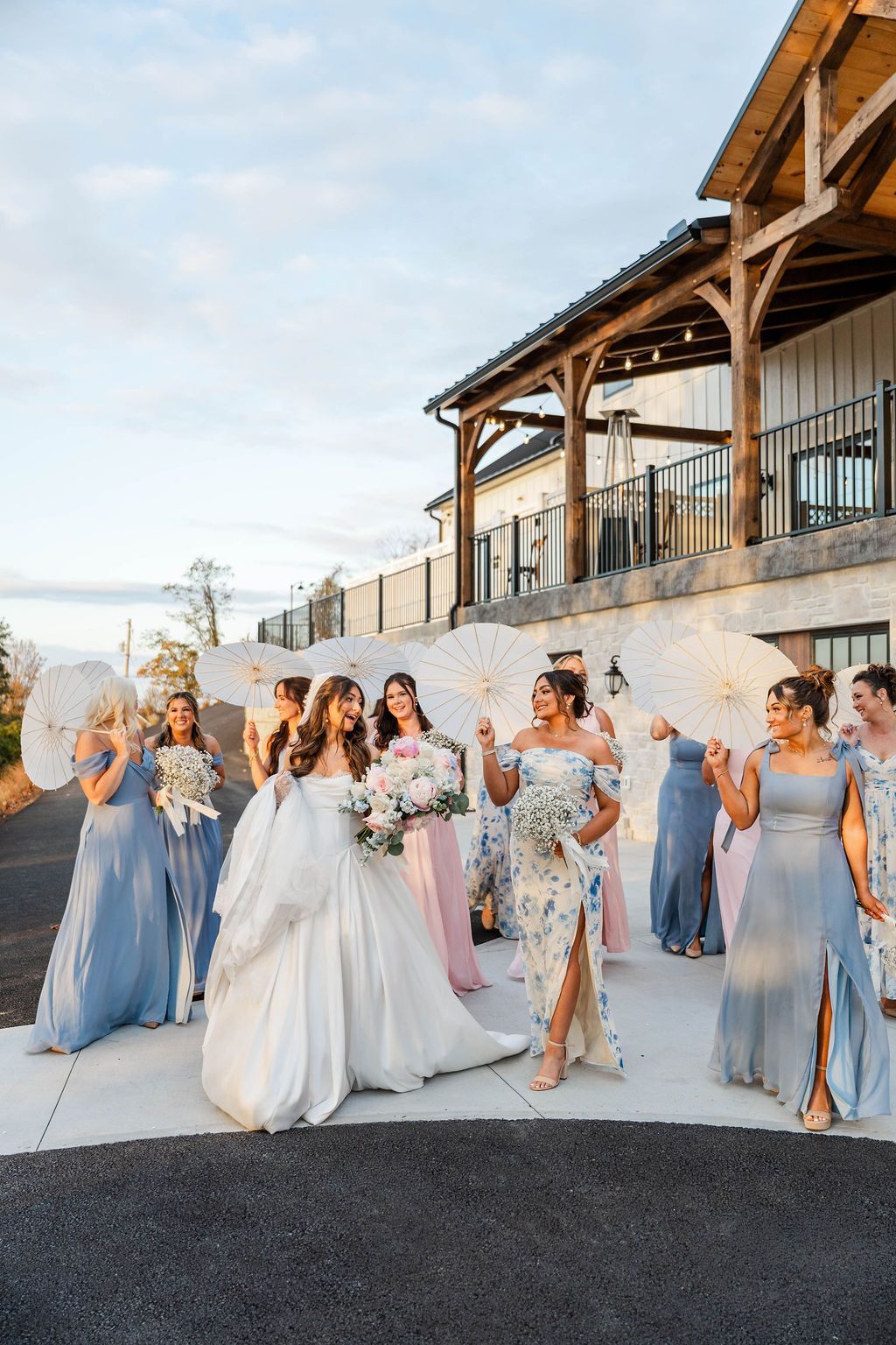 Bridal party in blue dresses and white gowns walking outdoors beside a wooden building at sunset