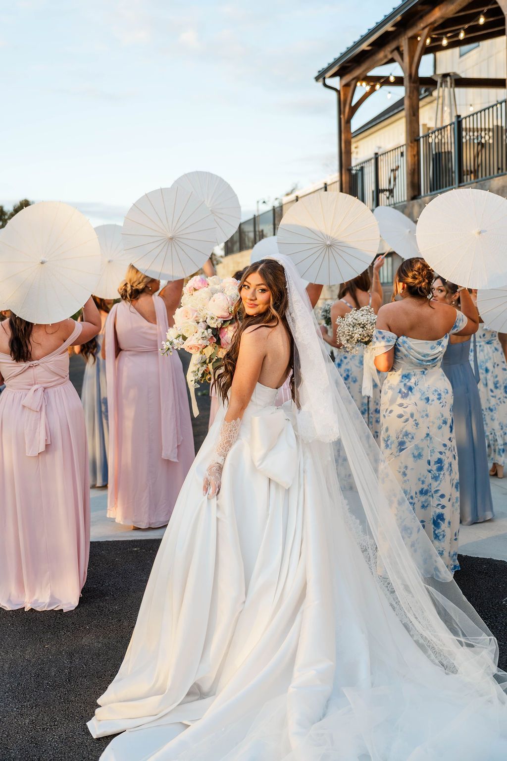 Bride in white gown posing with women holding white parasols outdoors at a celebration