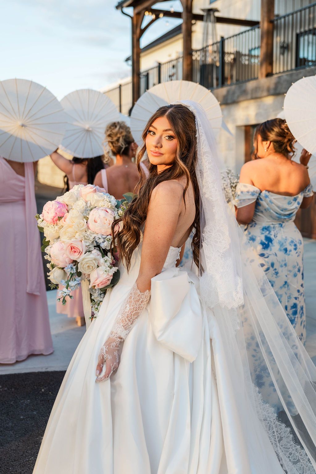 Bride in a white gown and veil holding a pastel bouquet, standing outdoors at sunset with guests behind her.