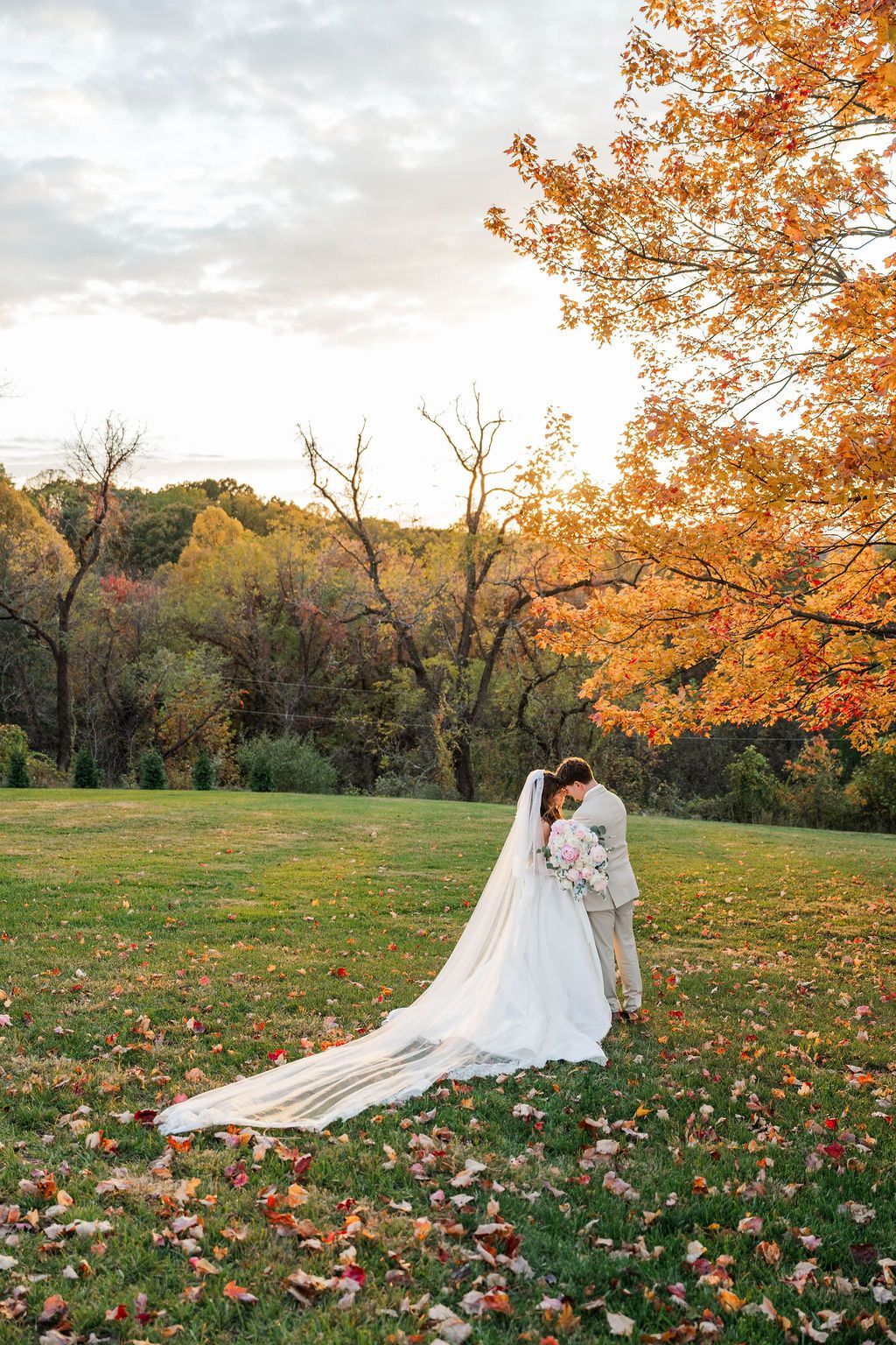 Bride and groom embracing in a grassy field with autumn trees and a long white veil trailing behind them