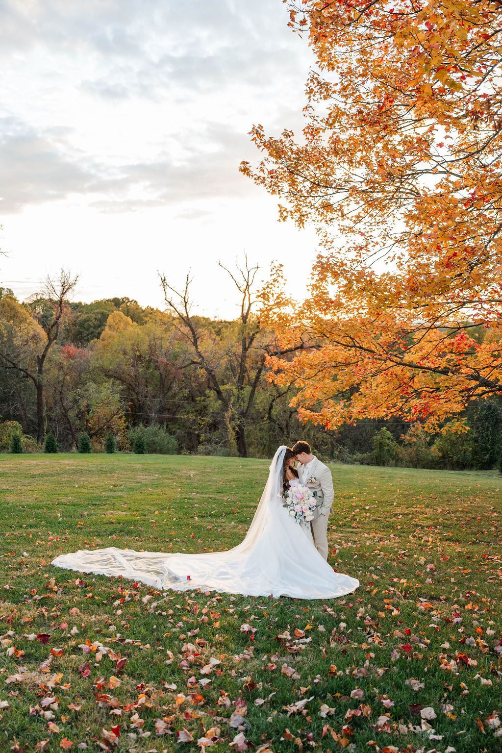 Bride in white gown and long veil standing in an autumn field beside an orange-leaved tree