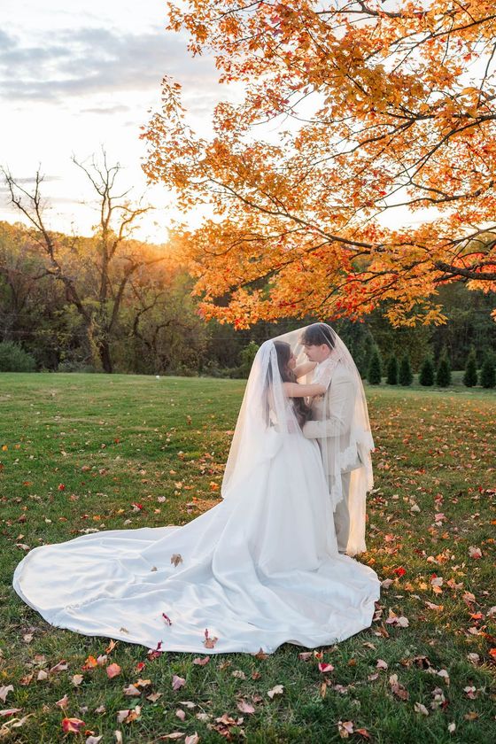 Bride and groom embracing in a park under orange autumn leaves, with a long white veil and train