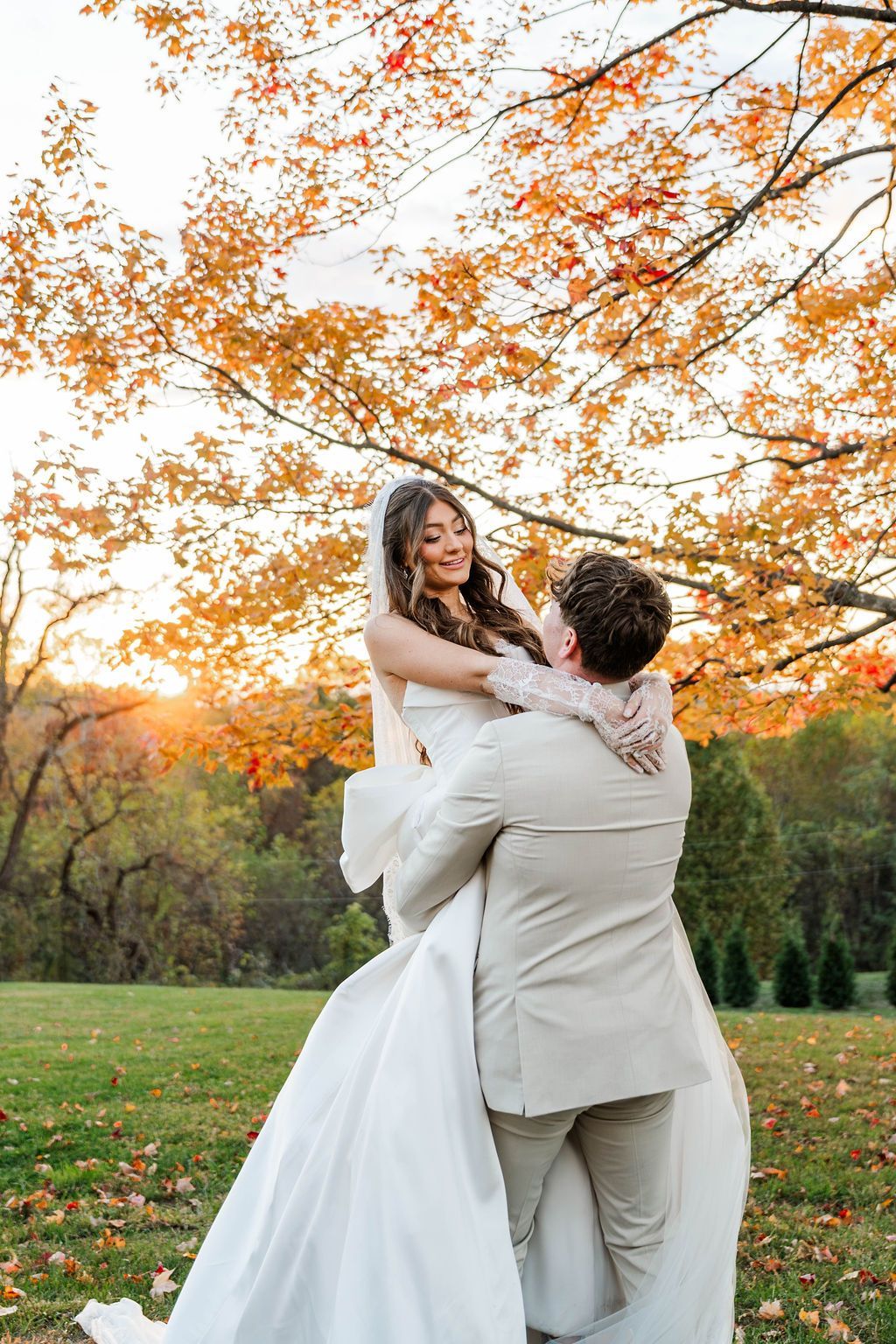 Couple in white wedding attire embracing outdoors beneath orange autumn branches