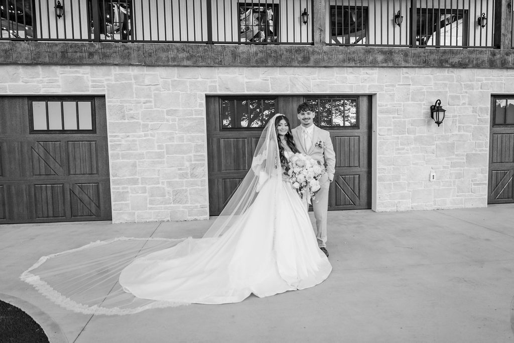Bride and groom posing in wedding attire beside a stone building, with the bride’s long veil and train flowing behind them