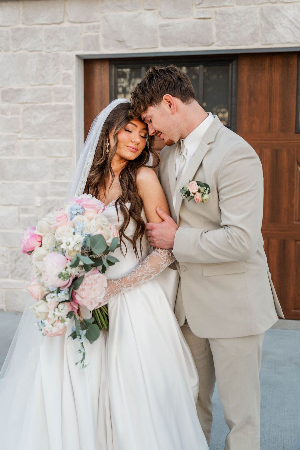 Bride and groom embracing by wooden doors, holding a pastel floral bouquet.