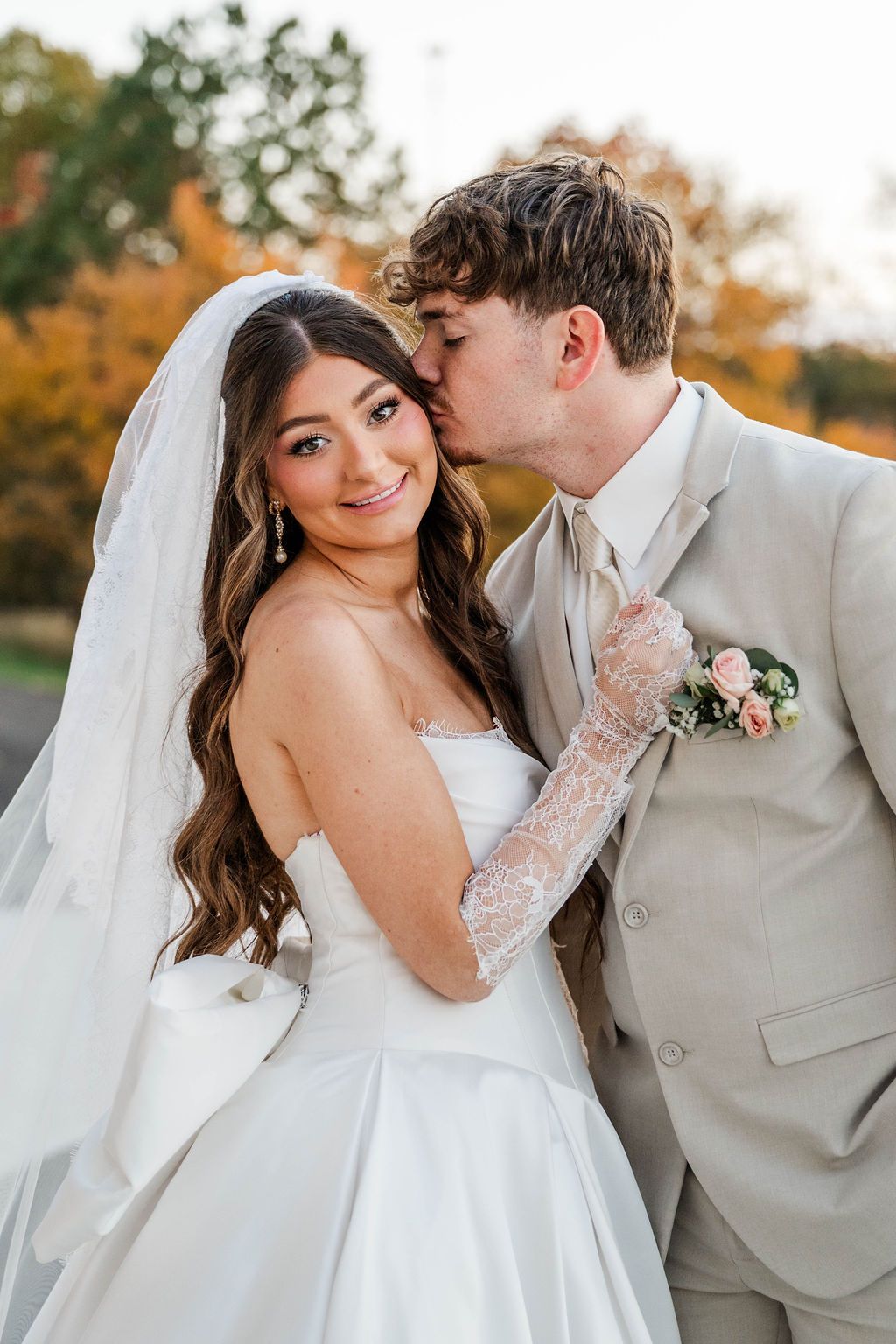 Bride and groom sharing a kiss outdoors, the bride in a white gown and lace gloves, the groom in a beige suit.