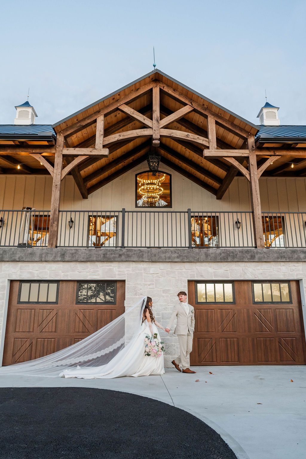 Bride and groom outside a rustic barn venue, bride holding a long veil at sunset