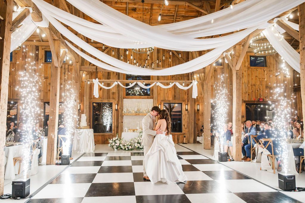 Bride and groom dance in a decorated hall with white drapes, string lights, and black-and-white checkered floor