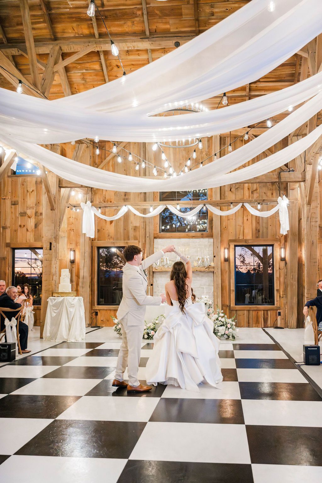 Bride and groom dancing in a rustic barn reception with black-and-white checkered floor and white drapery.