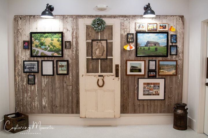 Rustic wall with framed photos, white door, and lamps. Wooden panels and decor create a farmhouse aesthetic.