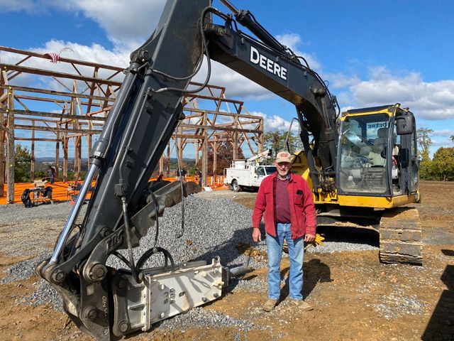 Man stands by a John Deere excavator at a construction site with a wooden structure in the background.