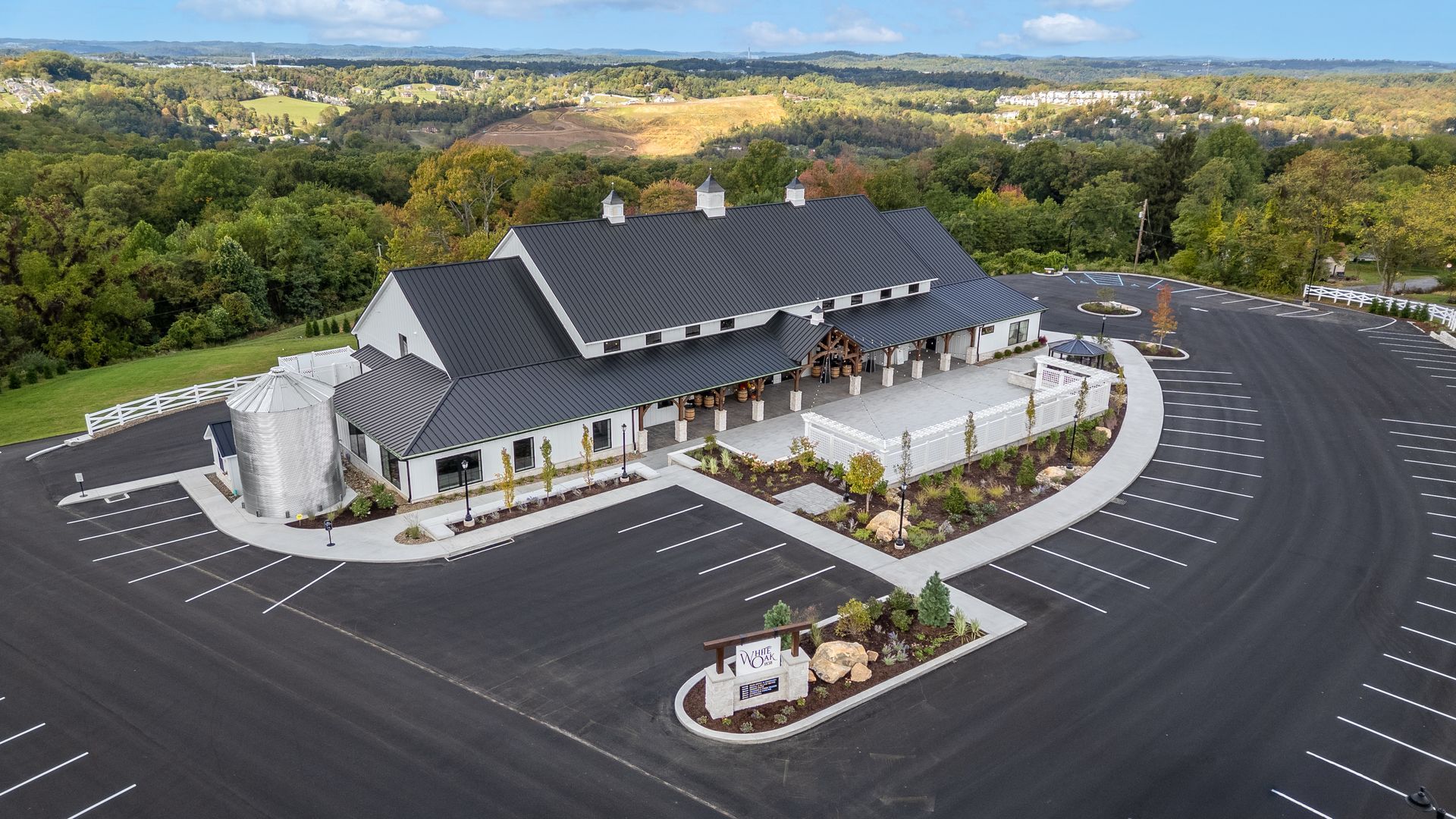 Aerial view of a large white building with a dark roof, surrounded by a mostly empty parking lot and greenery.