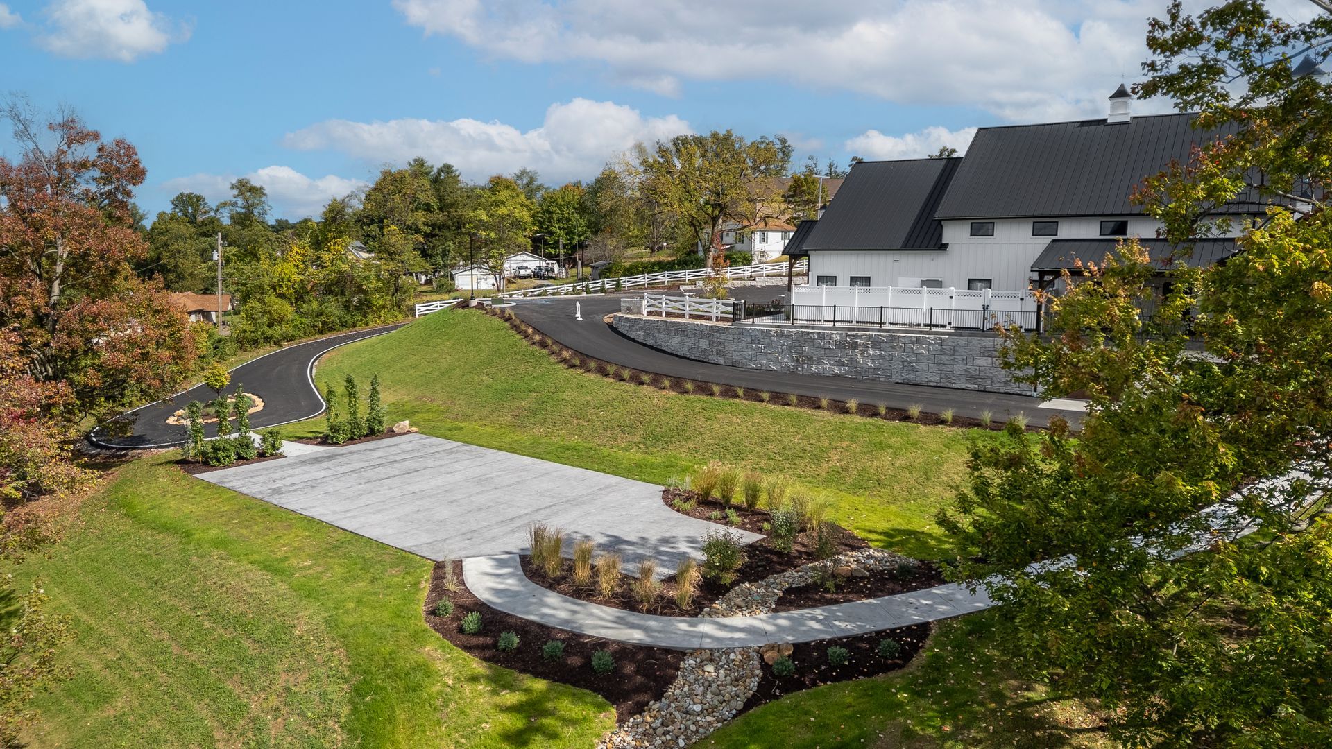 Curved driveway and landscaped lawn beside a white house under a blue sky with autumn trees.