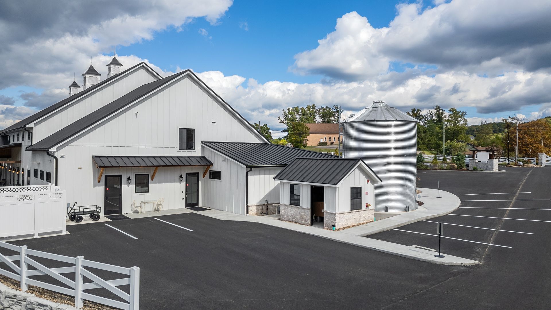 White building with black roof, striped silo, and parking lot under a cloudy sky