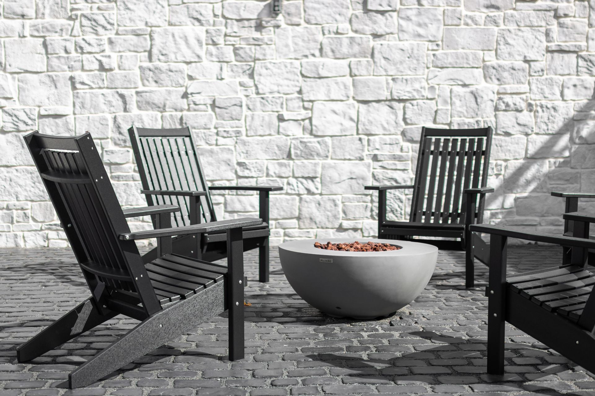 Outdoor patio with black chairs and a white fire bowl on a cobblestone surface against a white brick wall