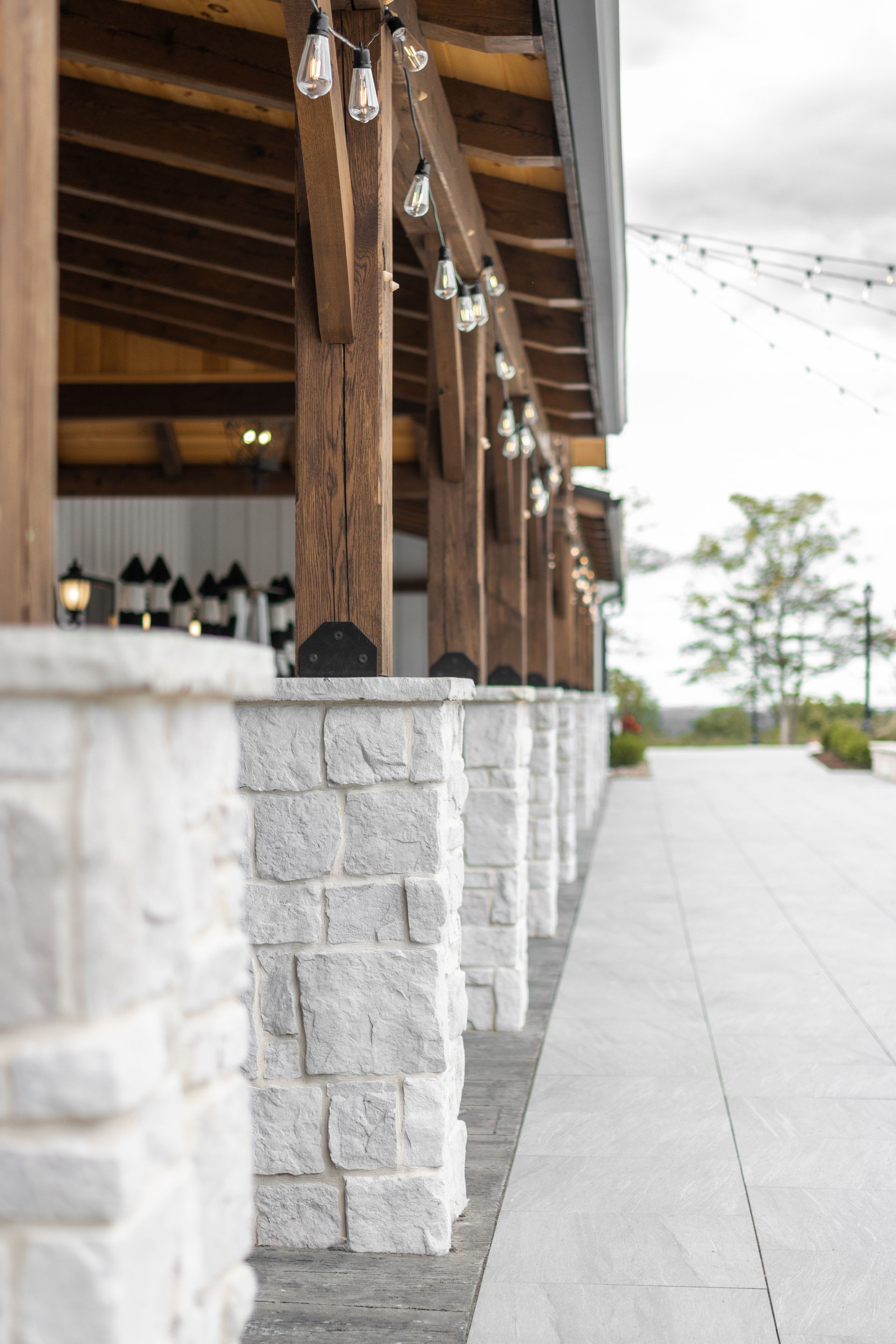 Covered stone walkway beside a rustic building with wooden posts and string lights, leading to trees in the distance
