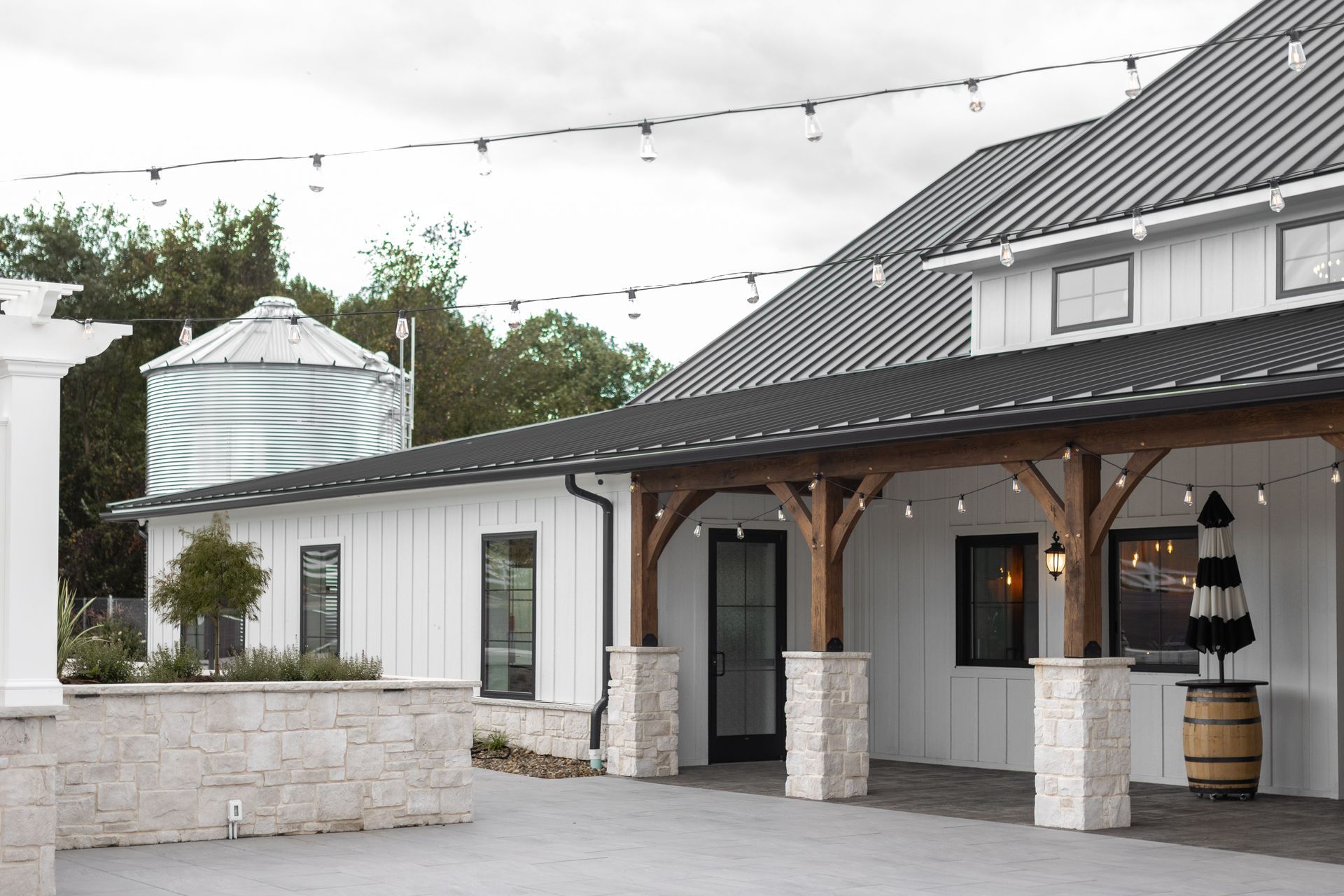 Outdoor patio of a white building with dark gray roof, stone pillars, and string lights at dusk