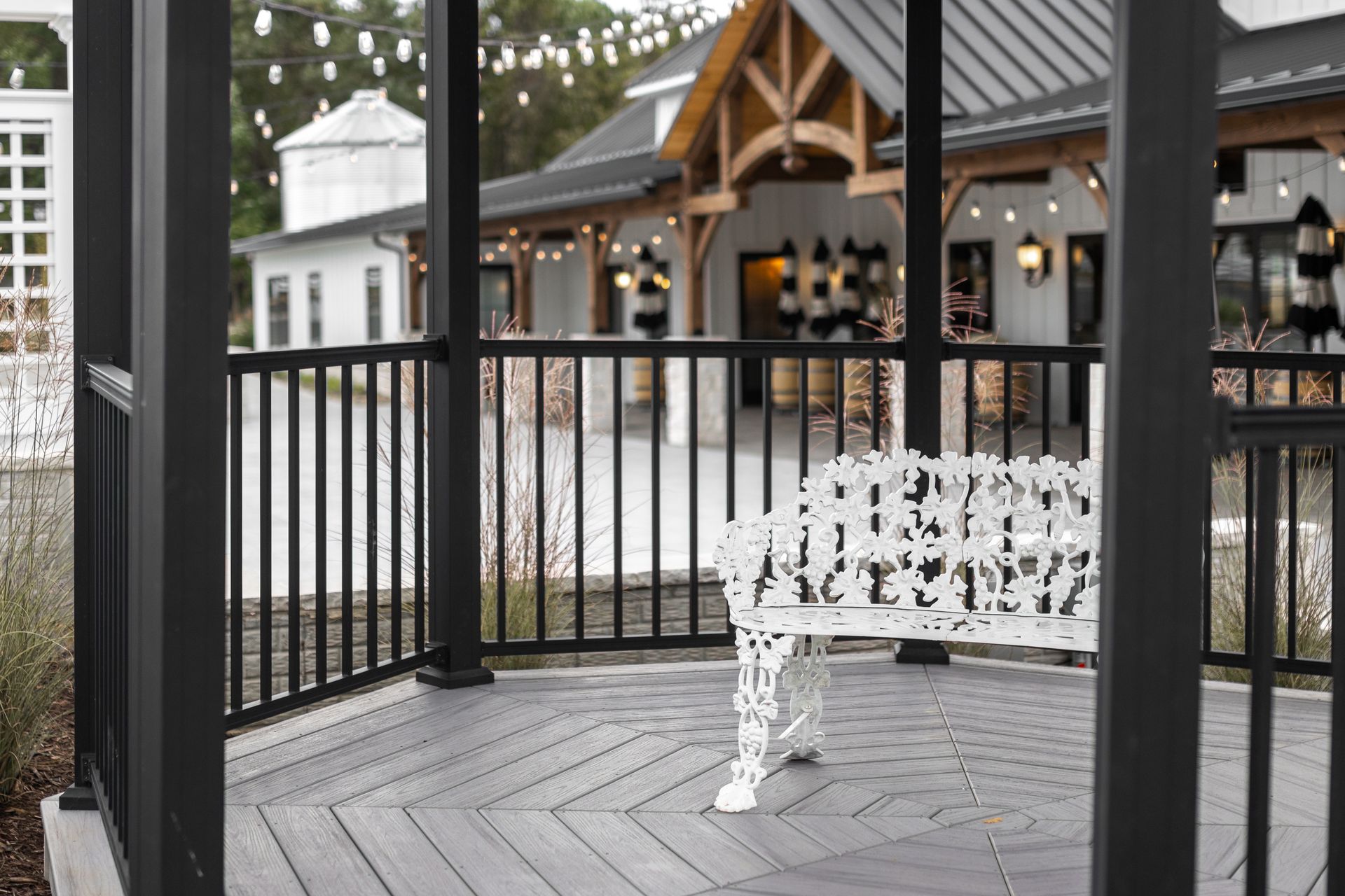 Black gazebo with white bench on a wooden deck beside a building with string lights