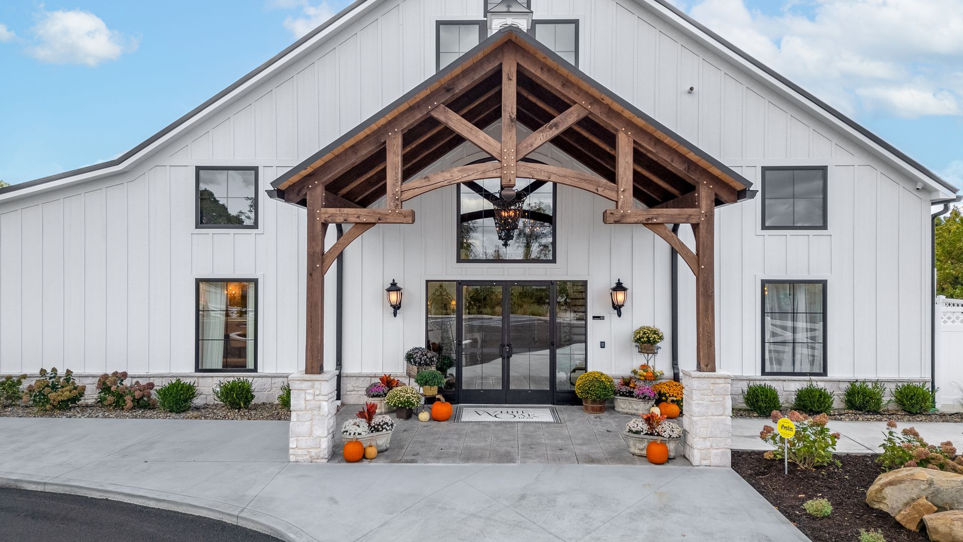 White barn-style building entrance with wood awning, glass doors, and autumn decorations by the walkway