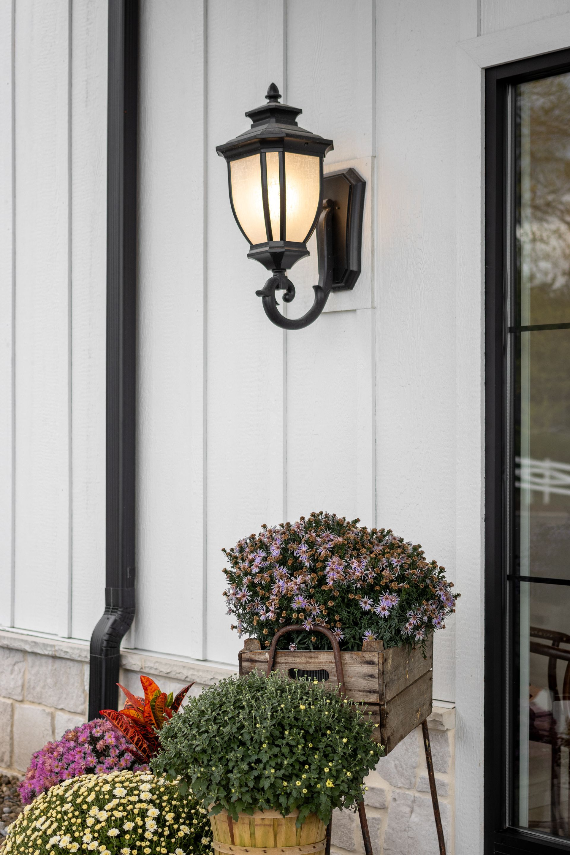 Front porch with a lit wall lantern beside potted flowers and a glass door