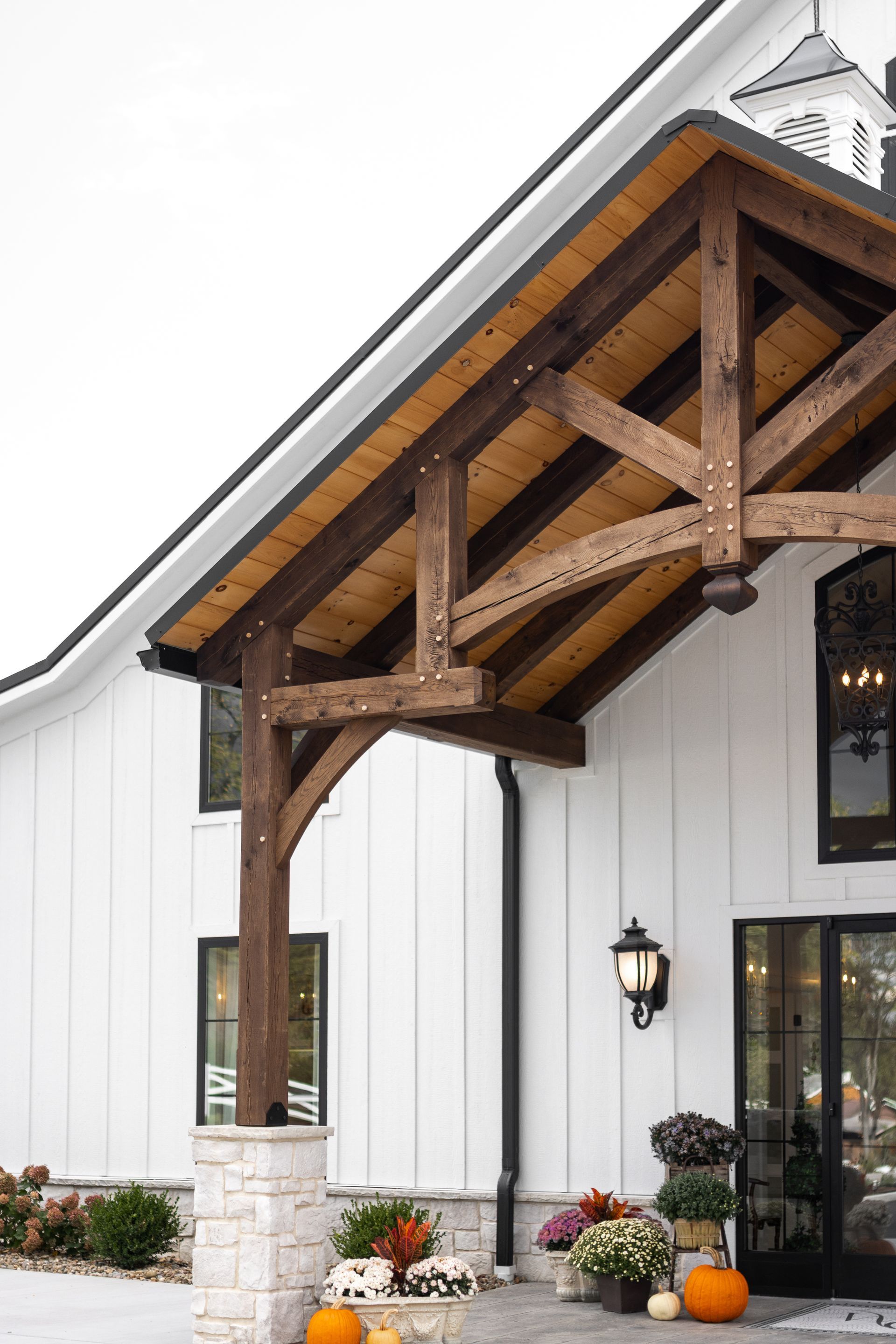 White farmhouse entry with rustic wooden porch roof, black lantern, and fall pumpkins by the door