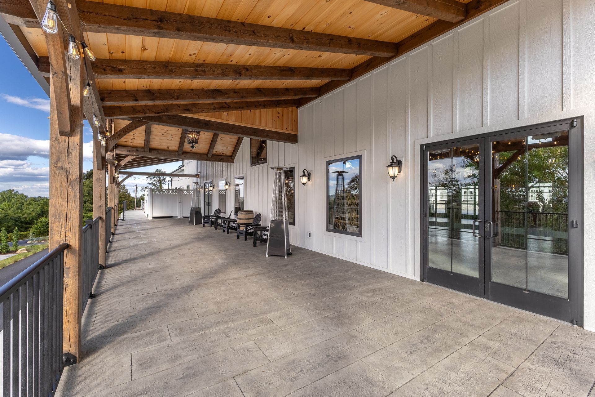 Covered outdoor patio with wood beams, concrete floor, chairs, and glass doors along a white wall