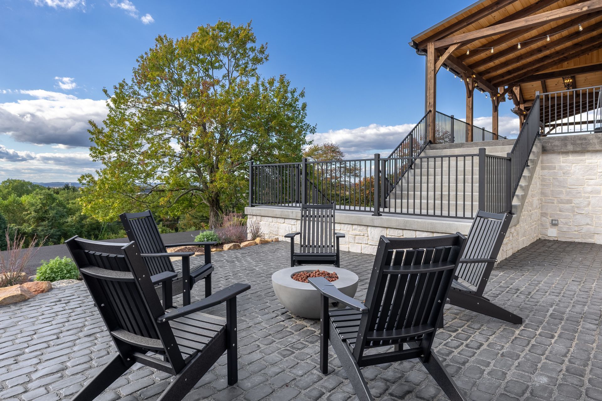 Outdoor patio with black chairs around a fire pit beside a modern house and tree, under a blue sky