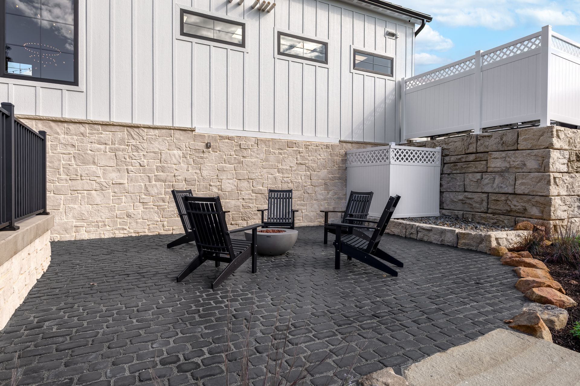 Patio with black chairs and table on stone pavers beside a white and stone house