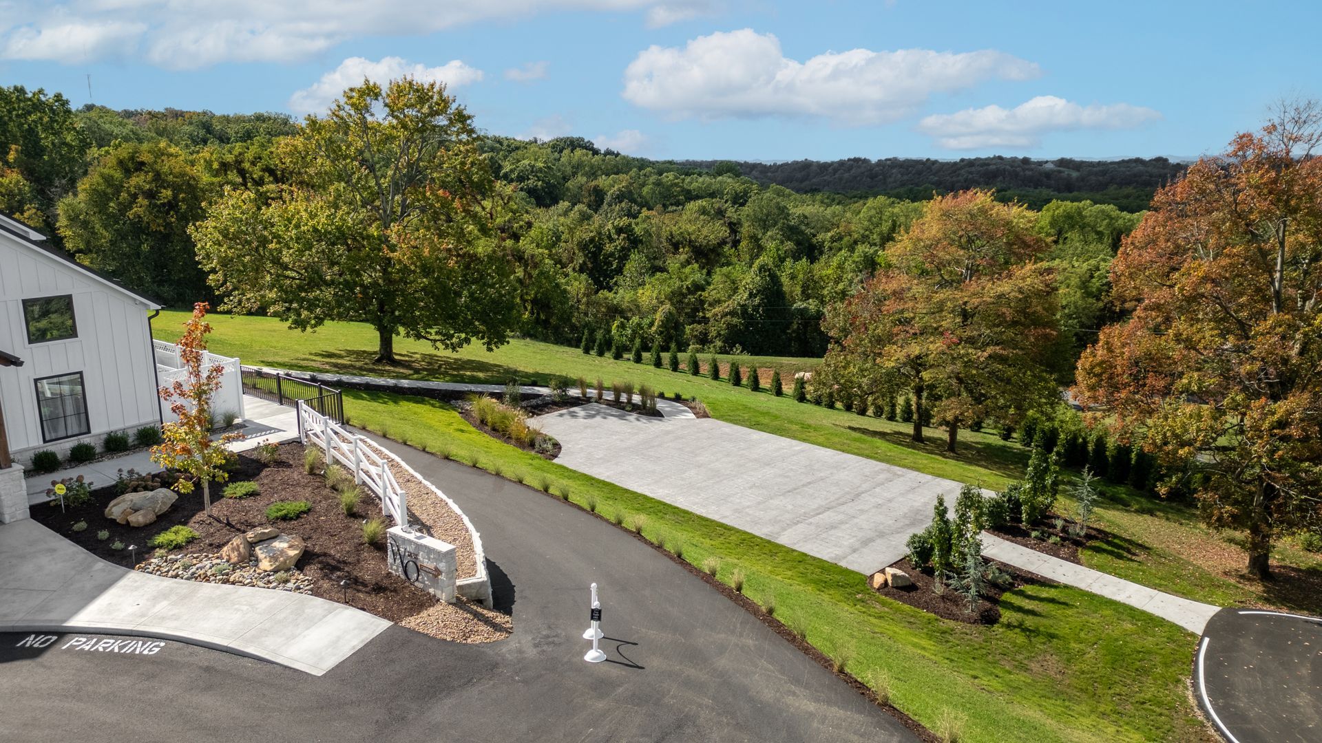 Rural driveway and house beside green lawns, trees, and hills under a clear sky