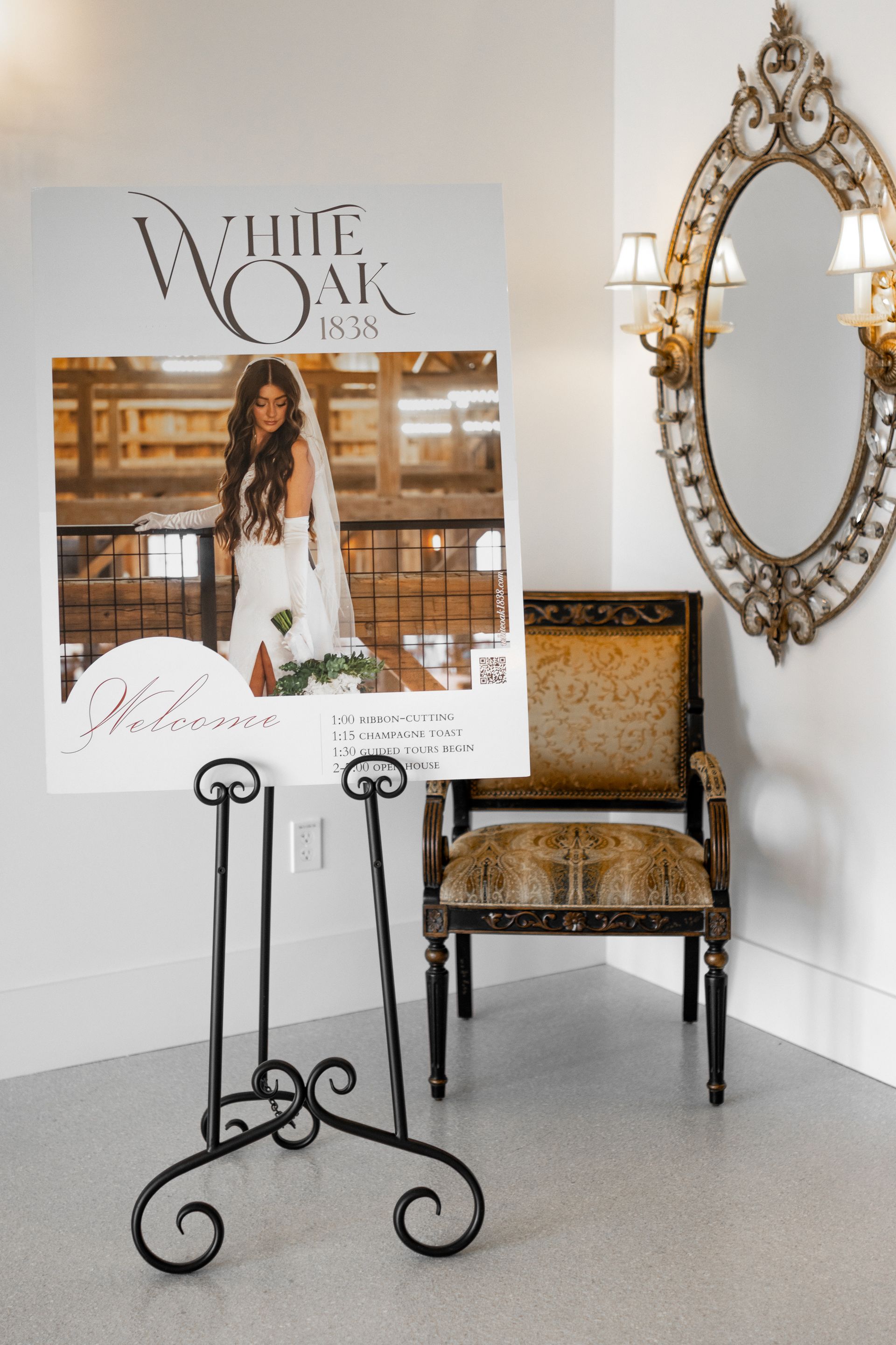 White easel with bridal poster beside ornate gold mirror and chair in a bright corner room