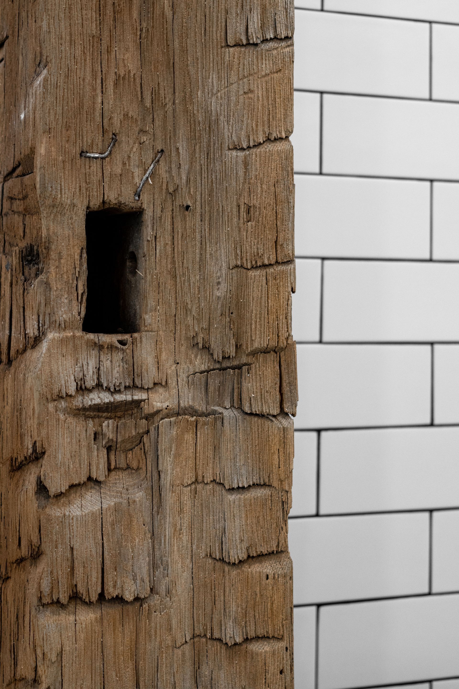 Close-up of a rough wooden beam beside a white tiled wall, with a dark rectangular opening in the wood