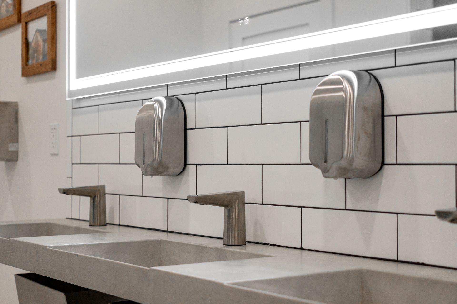 Modern restroom sink counter with white subway tile, mirrors, and stainless steel soap dispensers