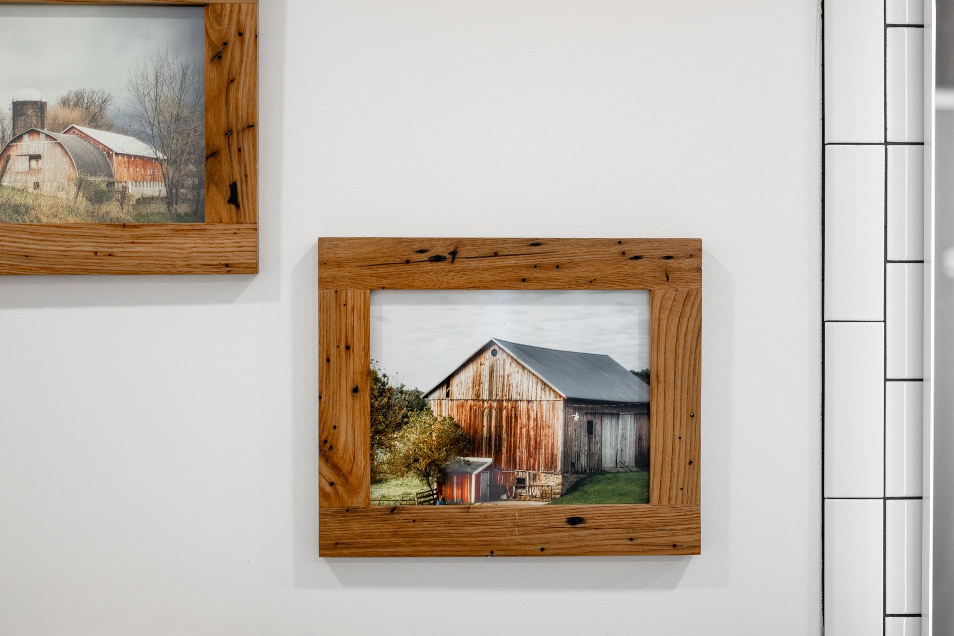 Framed rustic barn painting on a white wall beside a sliver of another framed artwork